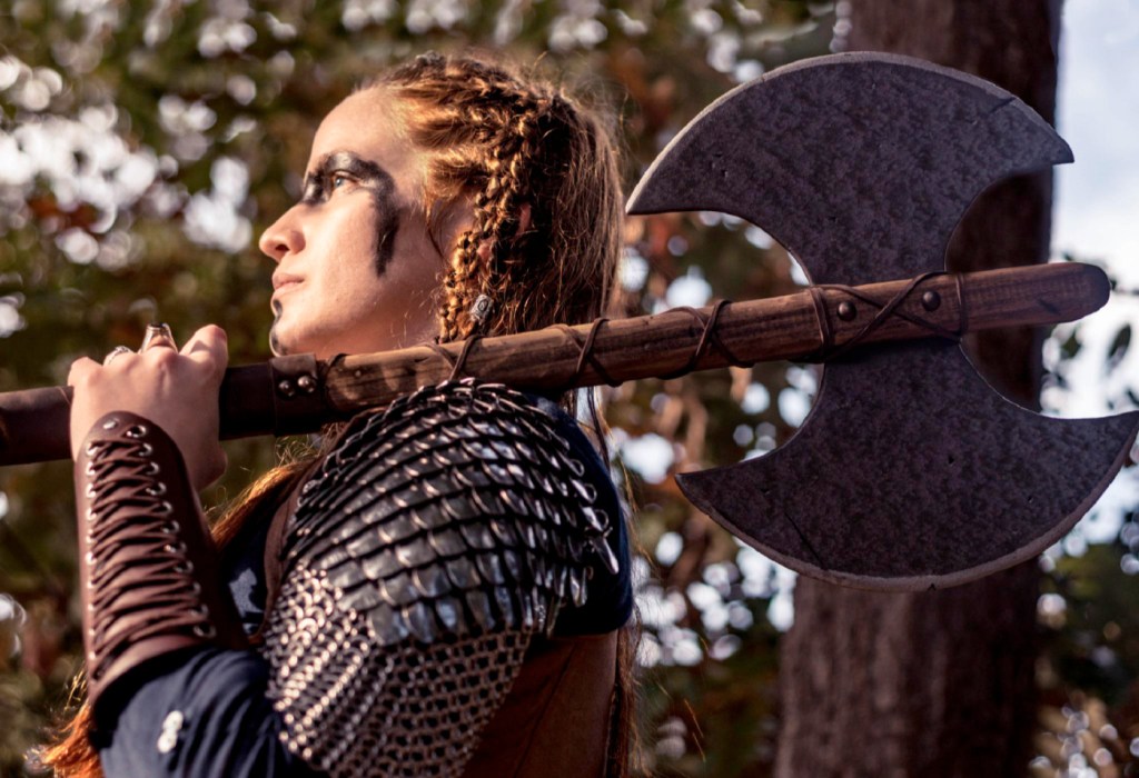A woman in armour holding an axe standing in a forest.